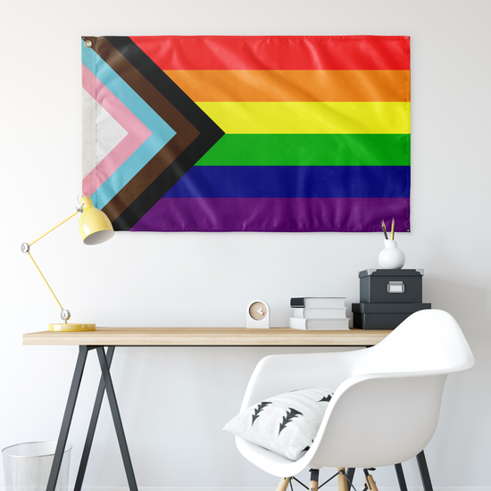 Image of a large rainbow progress flag on a wall. There is a modern desk and white modern chair below it.
