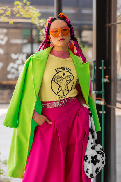 Photo of a fem-presenting person  wearing a Bella Canvas round-neck t-shirt and a bold neon outfit as she poses in the street. The Tshirt is yellow, with a black Sober for Satan graphic on the front.