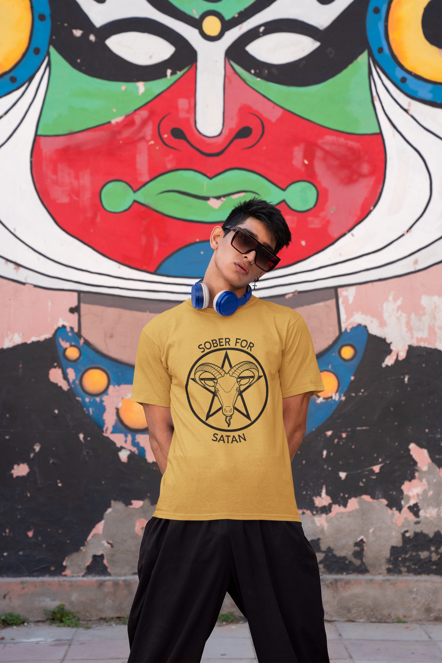 Photo of a young Indian man wearing a gold round-neck Gildan t-shirt while posing against a graffiti wall. The t-shirt has a black Sober for Satan graphic on the front.