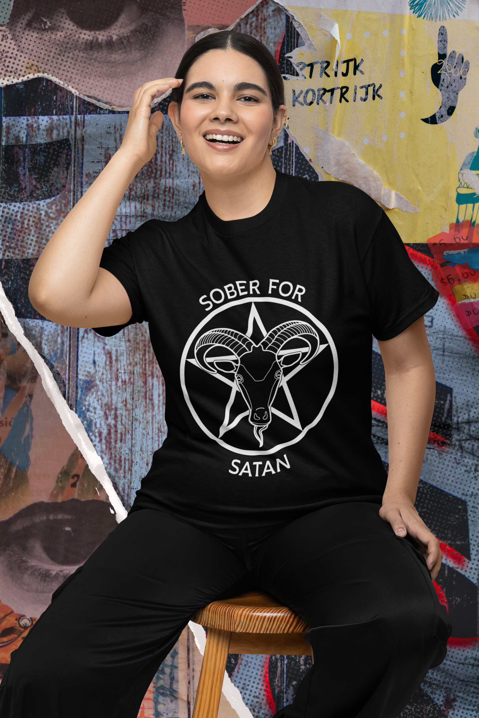 Photo of a happy person sitting on a stool, wearing black pants and a black tshirt with a white Sober for Satan graphic on the front.