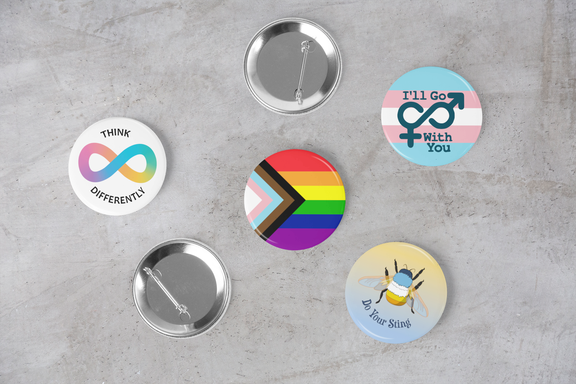 6 round pin-back buttons on a grey marble surface. Two are upside down, showing the pins. 1: Neurodiversity "think differently" 2: Raanbow progress 3: Transgender "I'll go with you" 4: Aroace V1 Bumblebee "Do your sting"