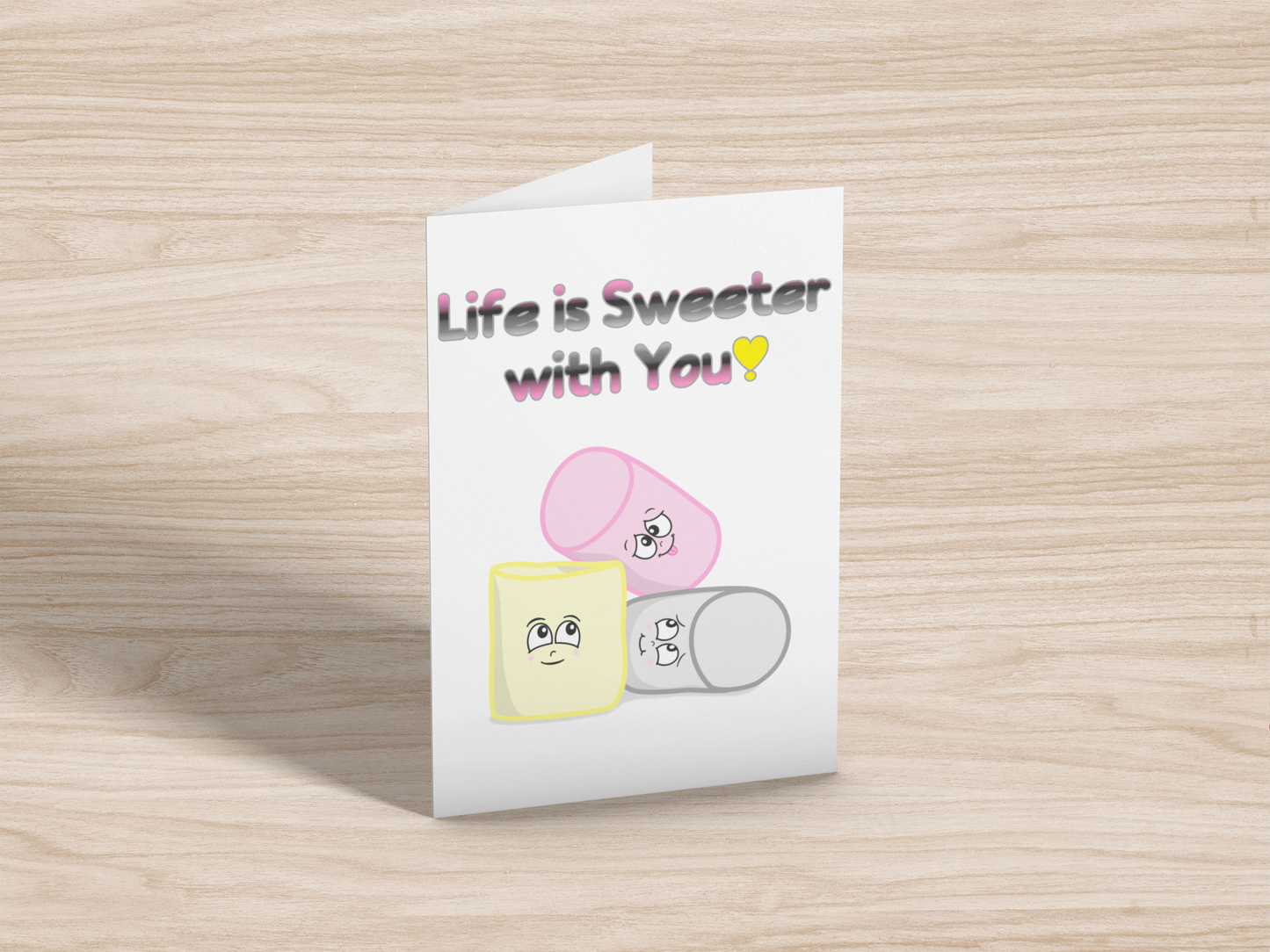 A greeting card on light wood background. The card has a white background. At the top is the text, "Life is Sweeter with You!" in the queerplatonic pride flag colours. Below are matching pink, yellow, and grey cartoon marshmallows with cute expressions. 